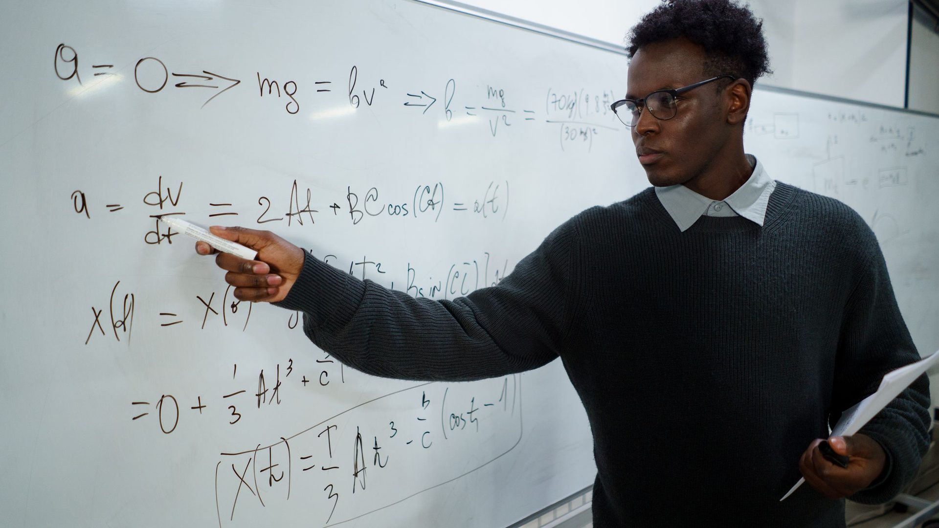 Jovem de óculos e camisola escura a apontar para uma derivada escrita num quadro branco