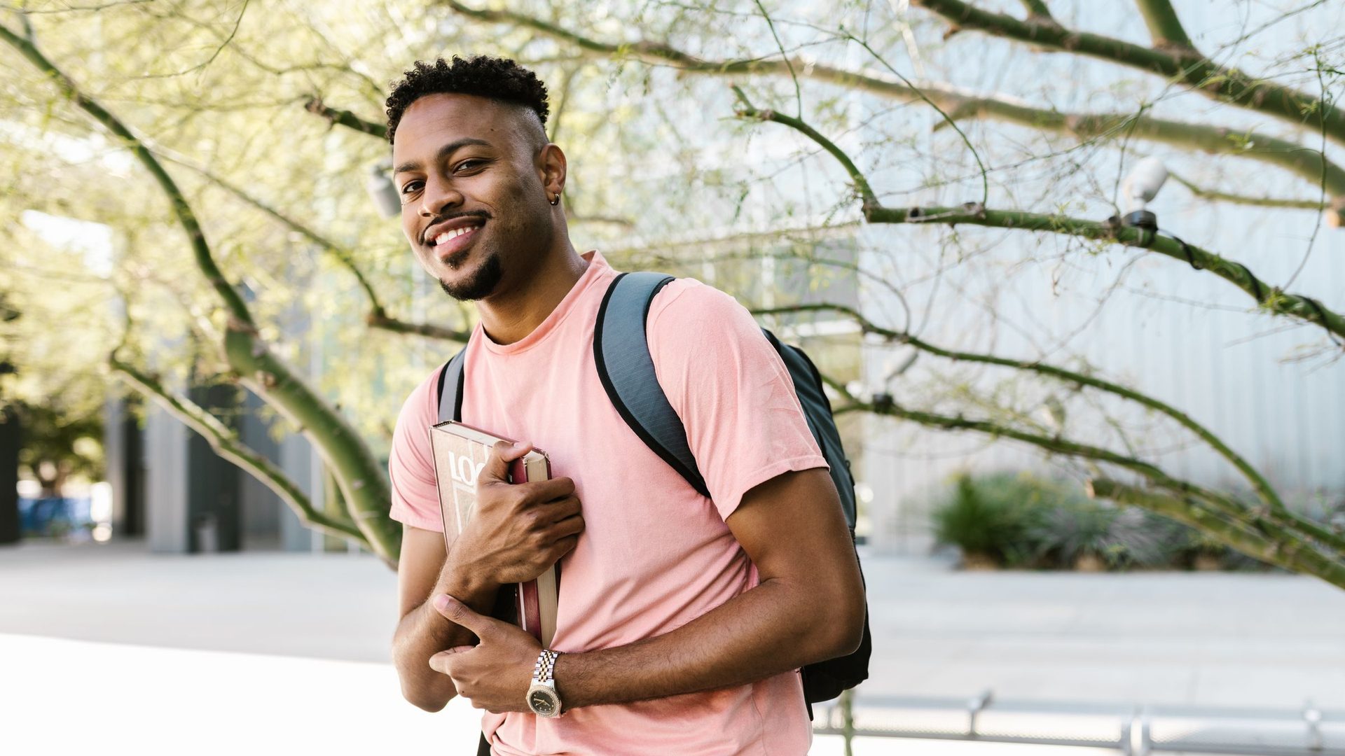 Estudante universitário sorridente de mochila às costas e livro na mão, num campus arborizado