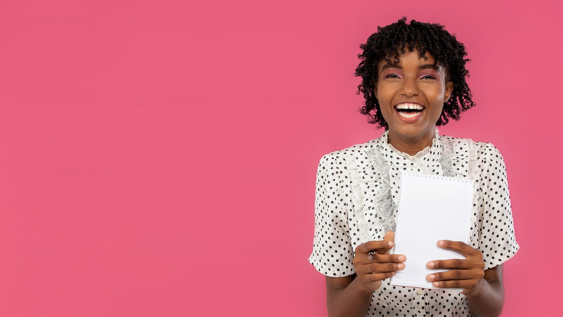 Jovem de blusa às bolinhas a rir com um caderno nas mãos, num fundo cor-de-rosa