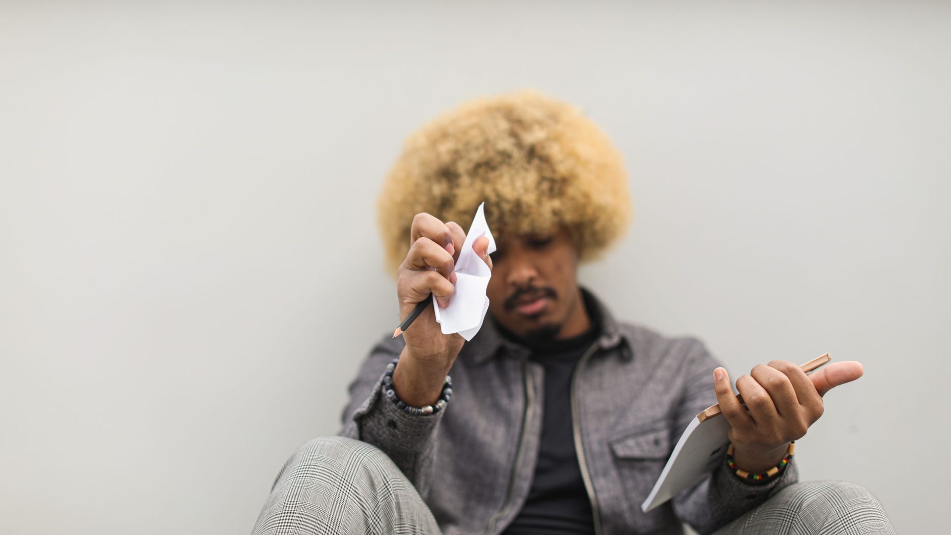Jovem de cabelo loiro encaracolado sentado no chão, frustrado, com uma folha amarrotada na mão e um caderno