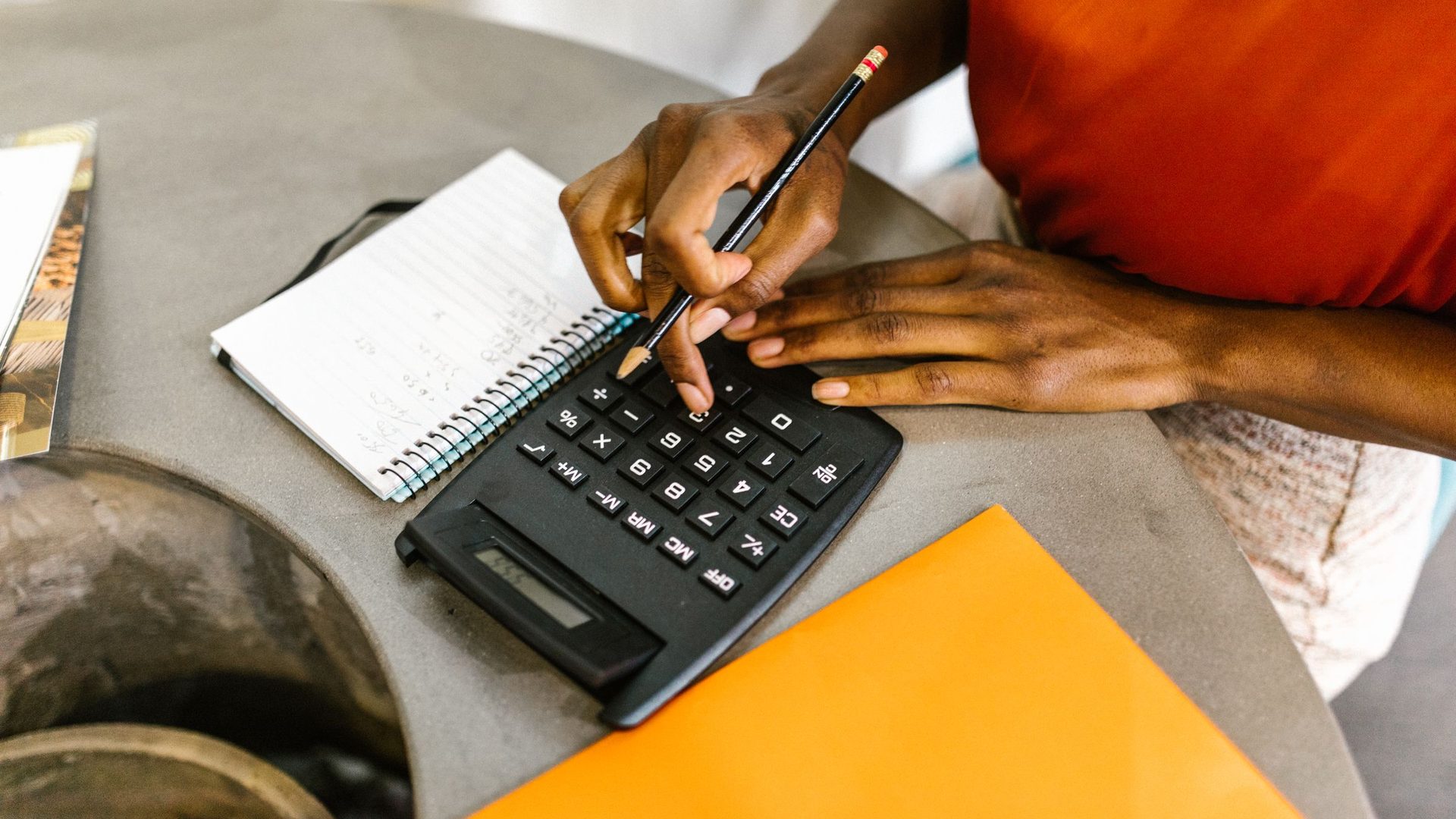 Mãos de uma estudante a fazer contas numa calculadora, com um caderno e uma pasta laranja na mesa