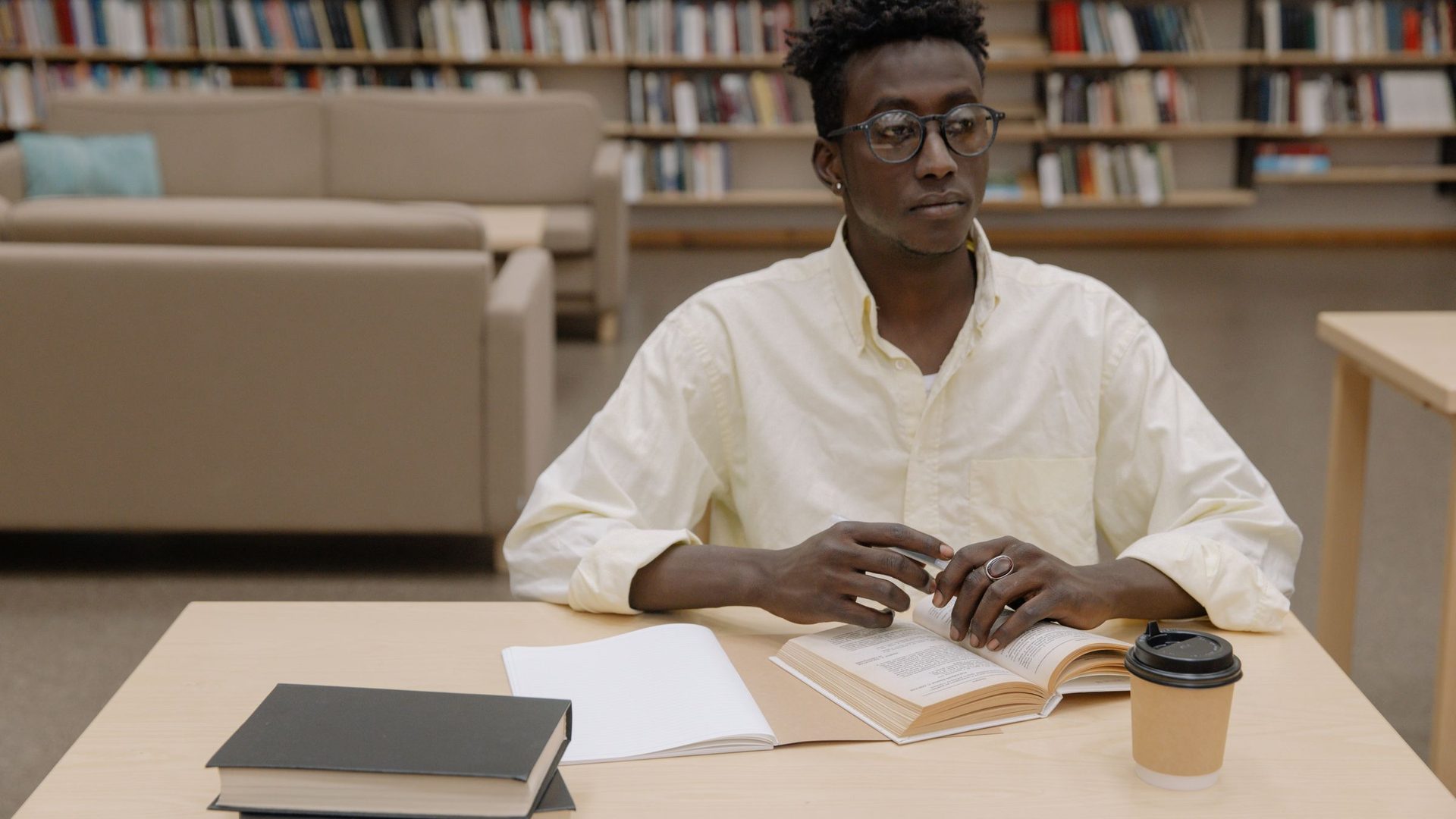 Jovem de óculos sentado a uma mesa de biblioteca, com um livro aberto e um café