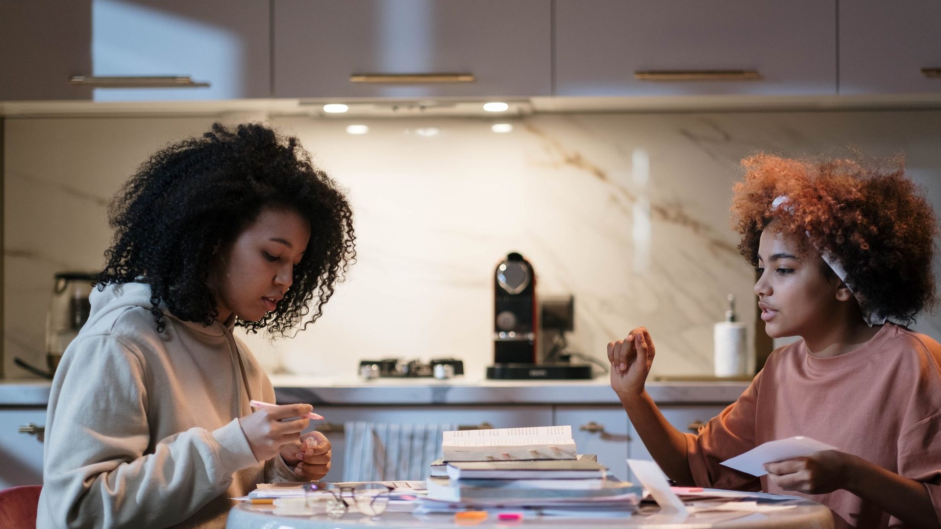 Duas jovens sentadas à mesa da cozinha com livros e cadernos, a estudar juntas