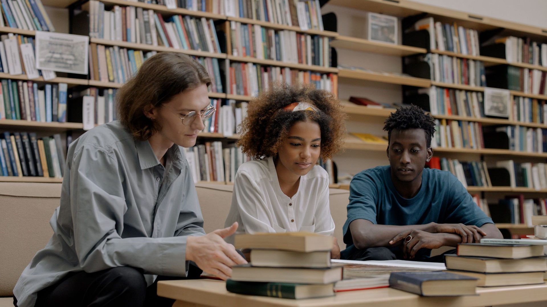 Três estudantes a estudar juntos numa biblioteca, com pilhas de livros na mesa