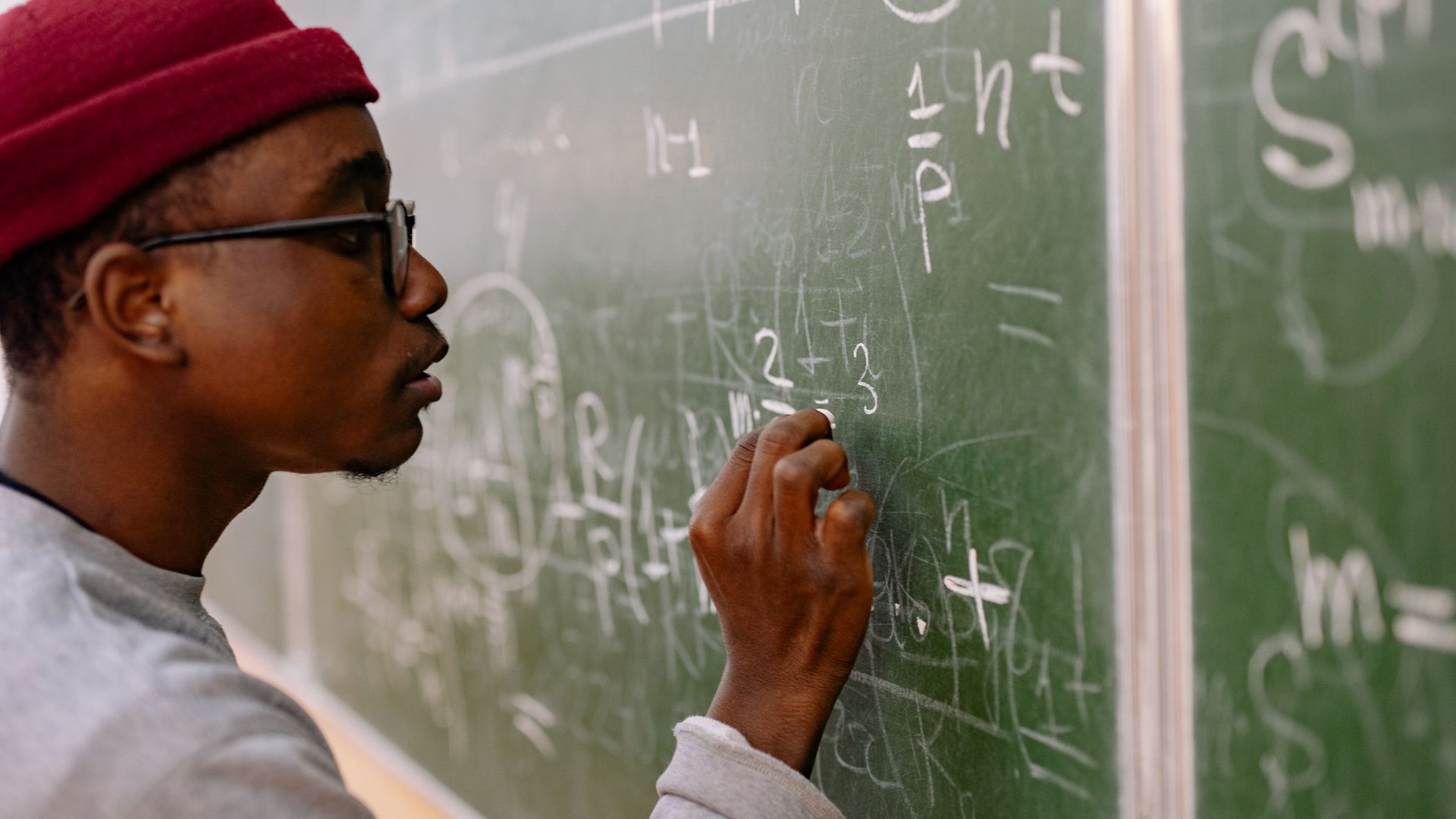 Estudante de óculos e gorro vermelho escreve fórmulas de matemática a giz num quadro verde