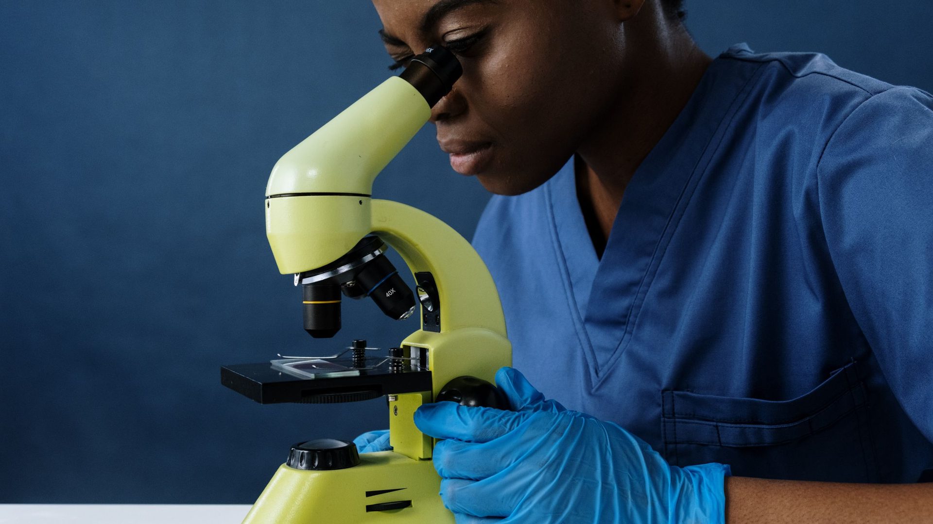 Técnica de saúde de farda azul e luvas a observar uma amostra ao microscópio num laboratório