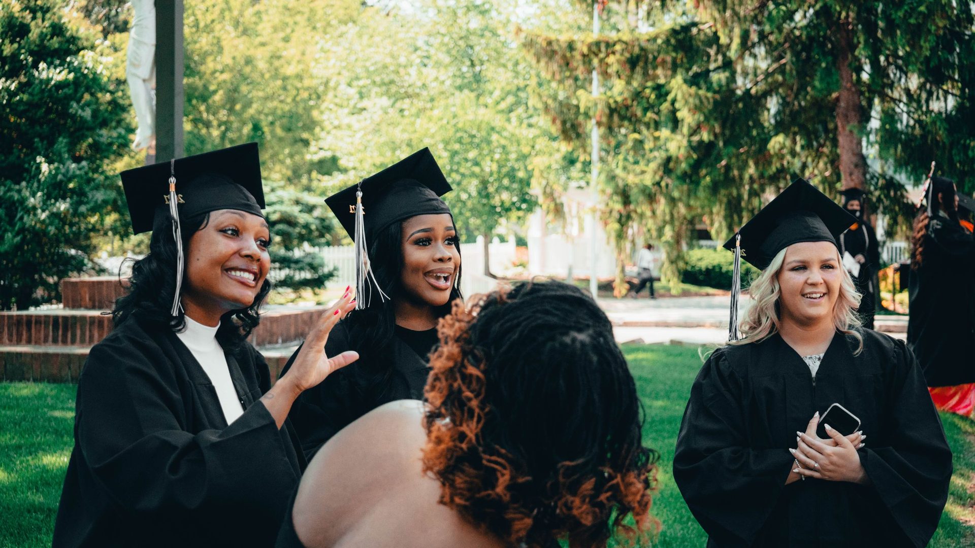 Grupo de jovens mulheres finalistas, de beca e capelo, a conversar e sorrir num jardim