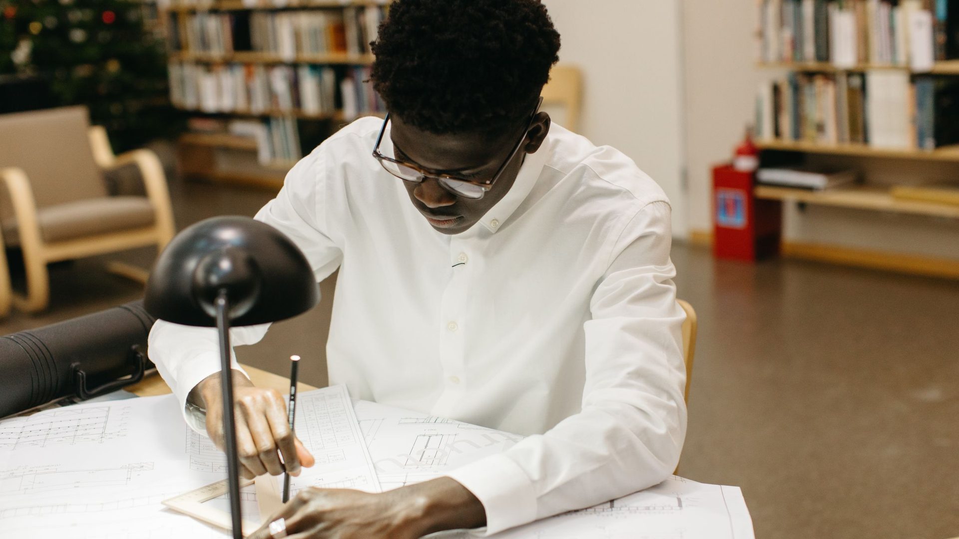 Jovem de camisa branca e óculos a desenhar uma planta técnica com lápis e régua numa mesa de biblioteca